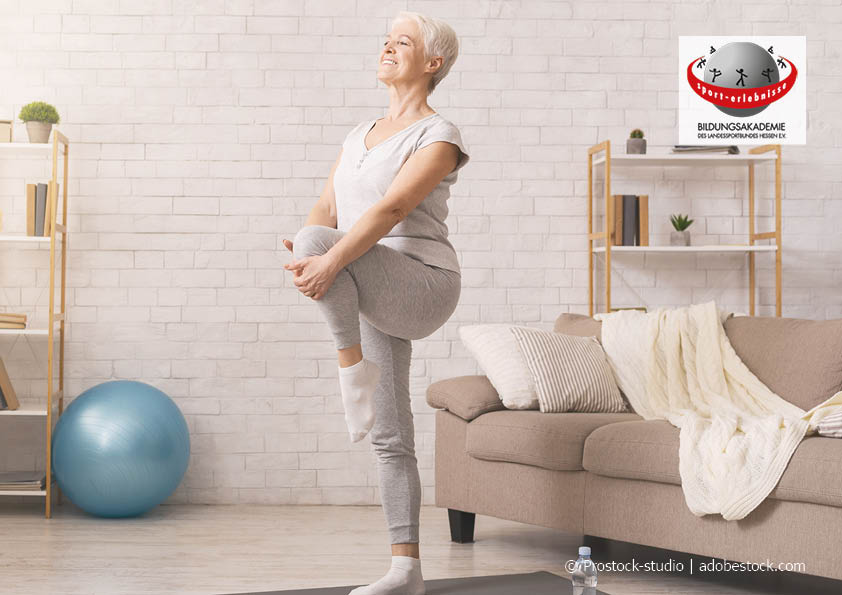 Ältere lächelnde Frau steht auf einem Bein auf Yogamatte in Wohnzimmer