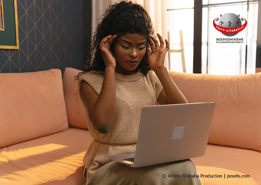 Eine junge Frau sitzt mit geschlossenen Augen auf dem Sofa und massiert die Schläfenvor einem Laptop beim Online Training Sehkraft stärken