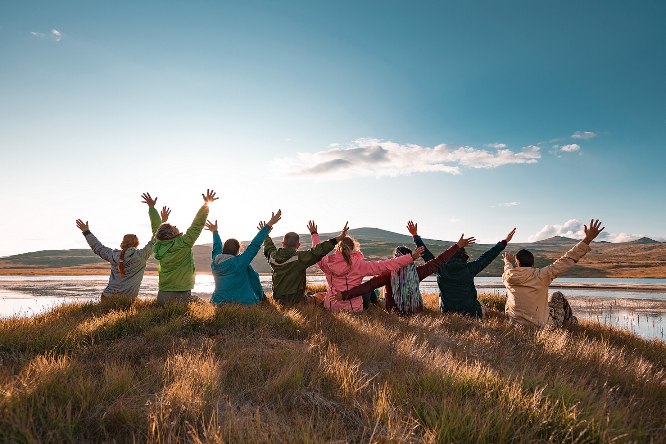 Eine Gruppe sportlicher Menschen sitzt gemeinsam auf einer Düne bei Sonnenuntergang und hebt fröhlich die Hände in die Höhe Freizeit Sport Fitness Übungsleiter Fortbildung Lizenz